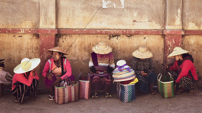A group of women sitting by concrete wall, selling items in baskets